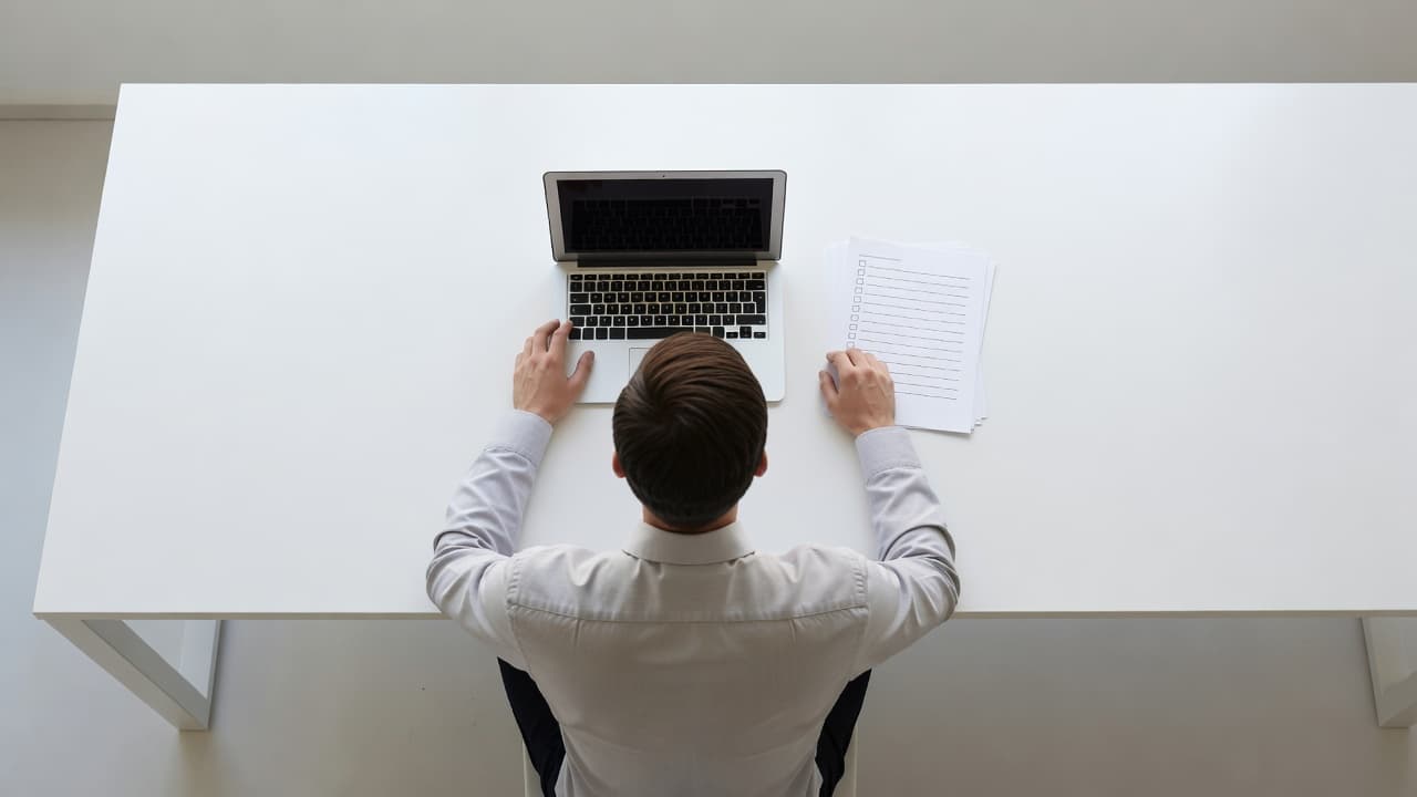 A person sits at a white minimalist desk from a top-down view, reviewing a clean paper checklist next to an open laptop. Soft daylight illuminates the organized workspace and professional documents.